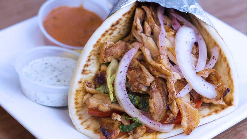 Gyro con dos salsas a los lados, una blanca y otra naranja, relleno de cebolla morada, carne y pimientos
