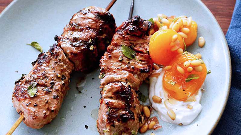 Carne en un pincho con una salsa blanca a un lado y dos albaricoques a los lados acompañada de piñones en un plato azul bebé 