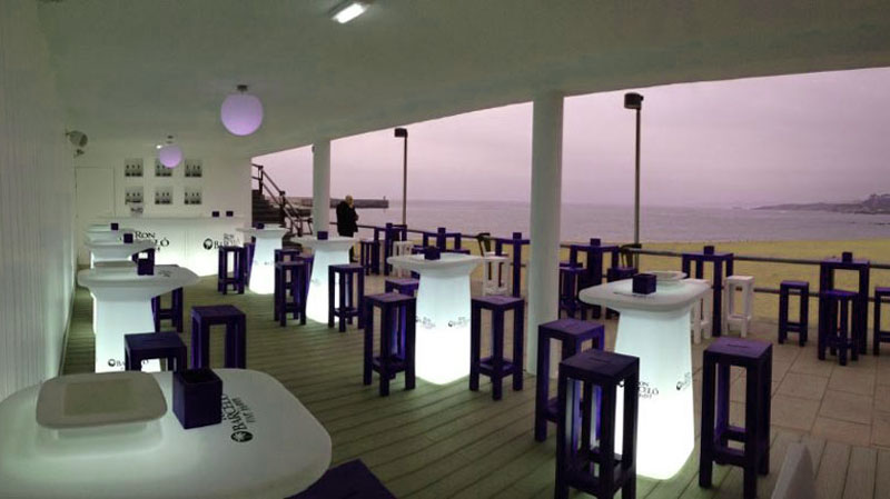 un chiringuito moderno con mesas blancas y taburetes azules delante de una playa