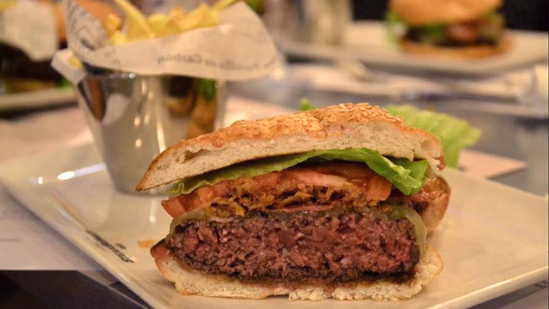 plato rectangular blanco con una hamburguesa partida por la mitad con un cuenco con patatas al fondo