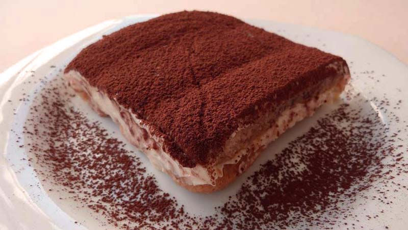 un trozo grande de tiramisú en un plato blanco con cacao espolvoreado