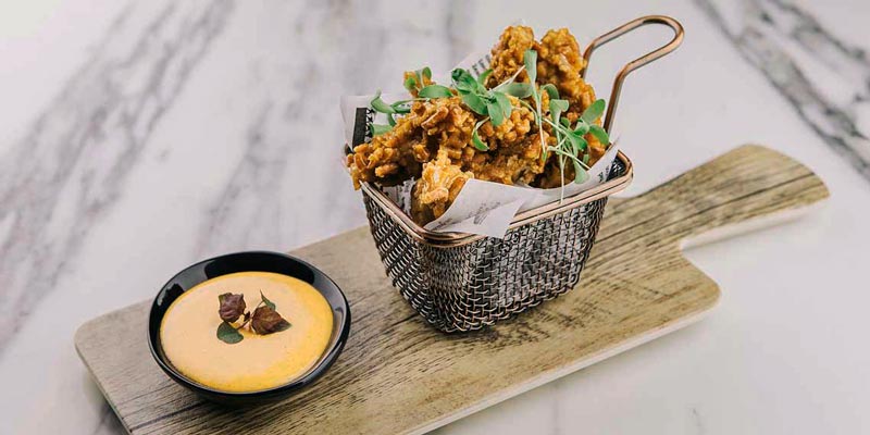 mesa de mármol con una tabla de madera que tiene una cesta con pollo frito y un bol negro con salsa