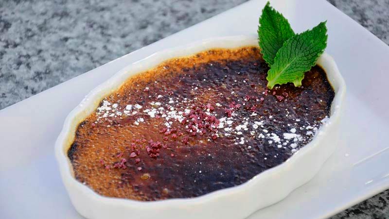 una crema en un recipiente de borde ondulado con hojas verdes que está sobre una bandeja que, a su vez está sobre una mesa de mármol