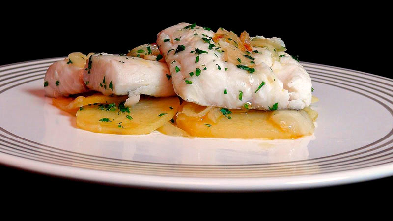 plato blanco con líneas que tiene en medio dos filetes de merluza con patatas y especias y sobre un fondo negro