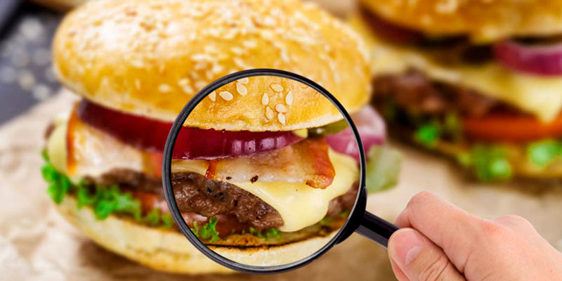una hamburguesa enfocada y otra detrás desenfocada, aparte hay una mano sosteniendo una lupa