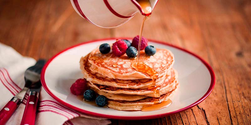mesa de madera con un plato blanco con el borde rosa y varias tortitas en el centro con frutos rojos a las cuales se les está echando sirope