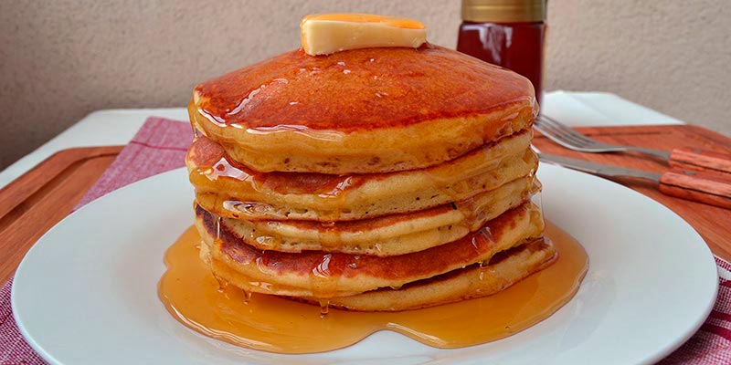 plato blanco con varias tortitas con sirope y que está sobre un mantel rojo y una bandeja de madera