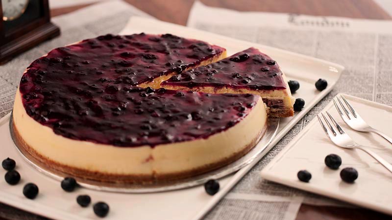 una tarta de queso completa en una bandeja blanca con arándanos y sobre una mesa que tiene papel de periódico