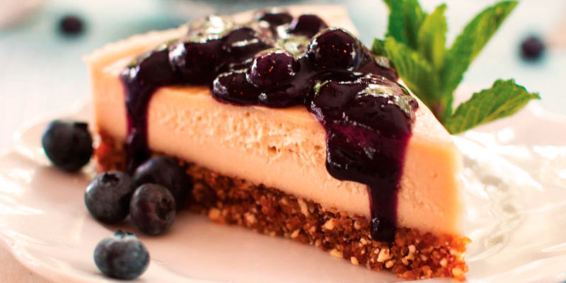 plato blanco con una porción de tarta de queso con salsa de arándanos y unas hojas verdes en la derecha
