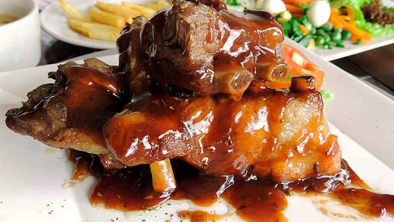 un plato blanco con varias piezas de costillas en salsa y con platos con verduras y patatas detrás