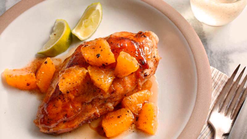 un plato redondo con una porción de pollo con salsa y trozos de naranja con un par de rodajas de limón