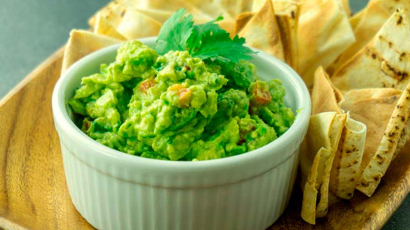bol blanco con guacamole rodeado de nachos y sobre un cuenco de madera