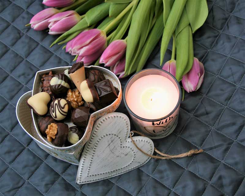 Ramo de tulipanes violetas junto a una caja en forma de corazón con chocolates dentro y una vela encendida.