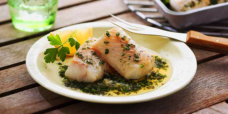 plato blanco con dos piezas de bacalao con una rodaja de limón y salsa verde, así como también un tenedor de dos púas. Todo ello sobre una mesa de madera. 