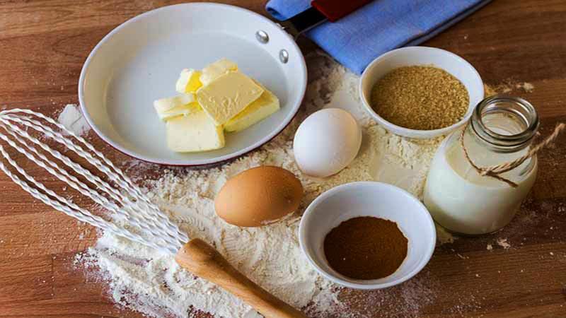 una mesa de madera con varillas, huevos, mantequilla, boles con ingredientes en polvo y una jarra con leche