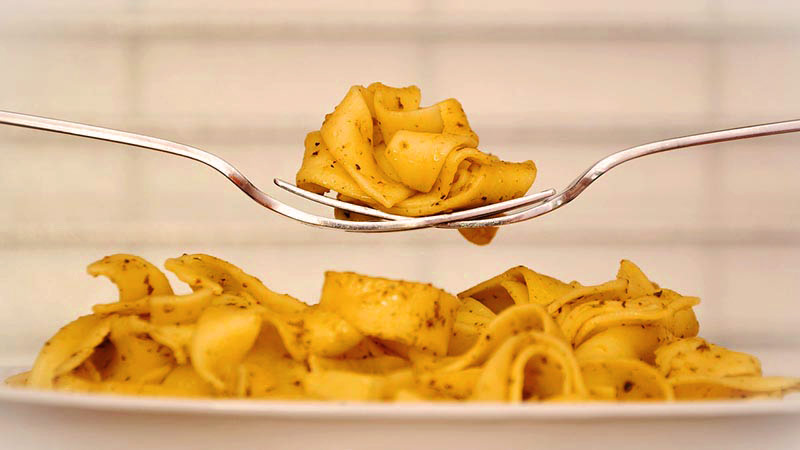 dos tenedores de perfil cruzados que sostienen fideos planos. Debajo tienen un plato con más fideos