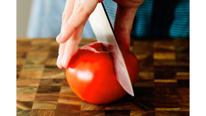 una mano sosteniendo un tomate y cortándolo con un cuchillo sobre una mesa de madera formada por cuadros