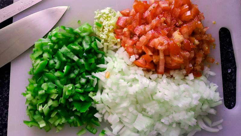 tabla de plástico con cebolla, ajo, tomate y pimiento verde picados con un cuchillo en la izquierda