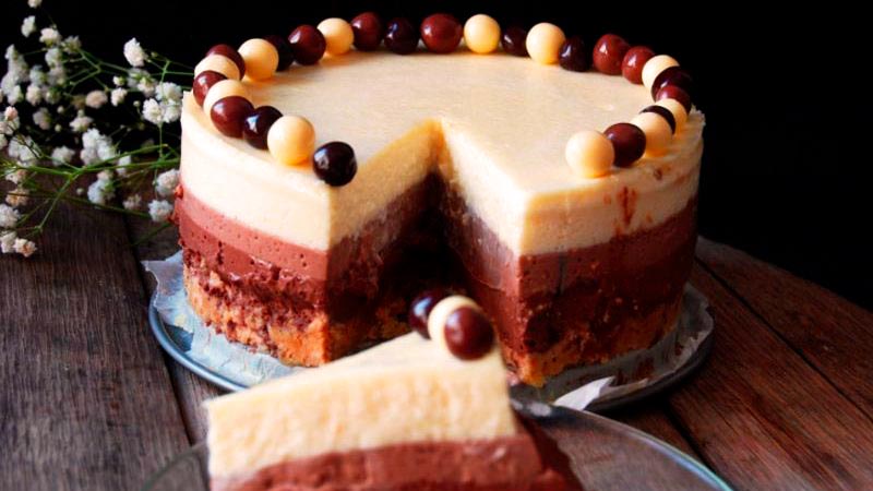 una tarta de diferentes chocolates, con un trozo partido y que está sobre una mesa de madera, con flores detrás y sobre un fondo negro