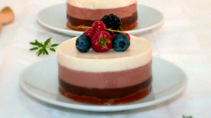 dos tartas redondas y pequeñas de tres chocolates que tienen frutos rojos encima y que están sobre unos platos y un fondo blancos