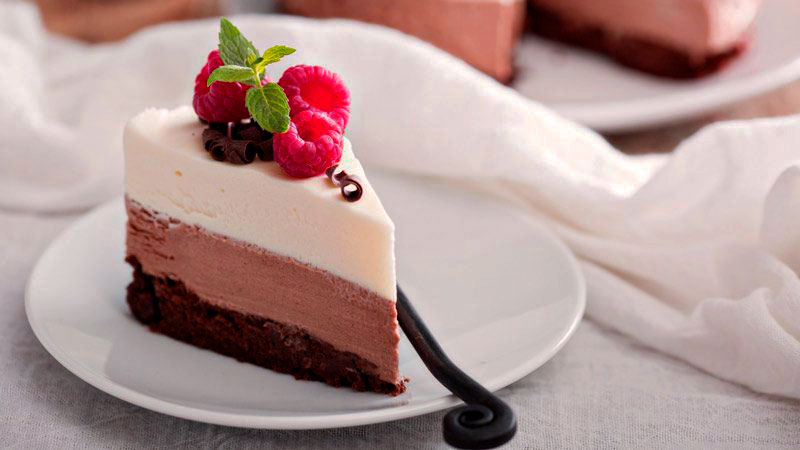 porción de una tarta de tres tipos de chocolate que está sobre un plato blanco y al lado de un mantel que tiene al fondo más tarta desenfadada