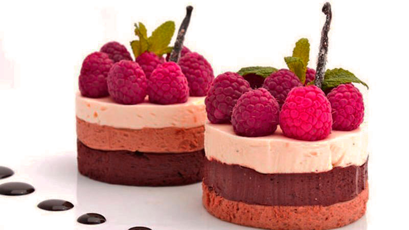 dos tartas pequeñas de tres chocolates con moras encima y sobre un fondo blanco con gotas de chocolate