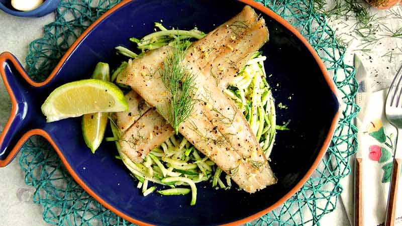 dos piezas de merluza sobre verdura con dos trozos de limón al lado y en un plato en forma de pescado azul