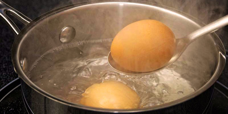 cacerola metálica con agua hirviendo y dos huevos, uno de ellos cogido con una cuchara metálica