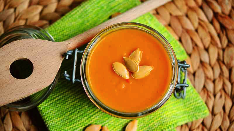 recipiente de cristal con crema hecha de calabaza con semillas de la misma y que está encima de un mantel verde con una espátula de madera al lado