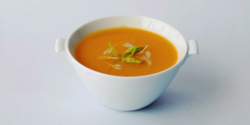 fondo blanco con un bol blanco lleno de crema anaranjada de calabaza con algunas hierbas en el centro