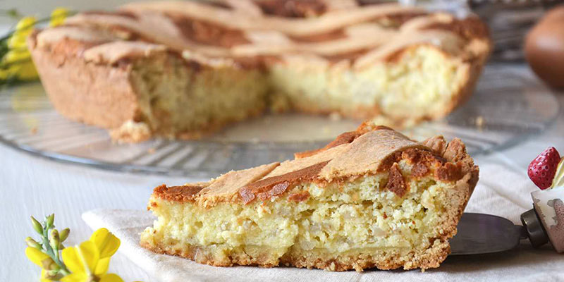 Una pastiera de napolitana sobre un plato de cristal y al lado, en una servilleta un trocito cortado. Alrededor flores amarillas a modo decorativo