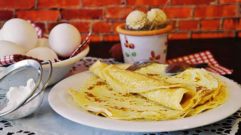 plato blanco con crepes que tiene un plato en la izquierda con huevos, una maceta al fondo y una pared de ladrillo también al fondo