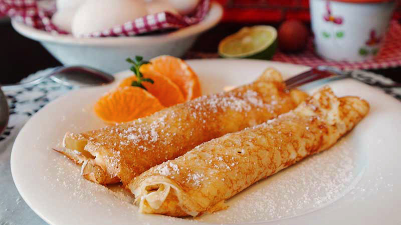 plato blanco con crepes que tienen gajos de naranja a un lado y un plato al fondo con huevos