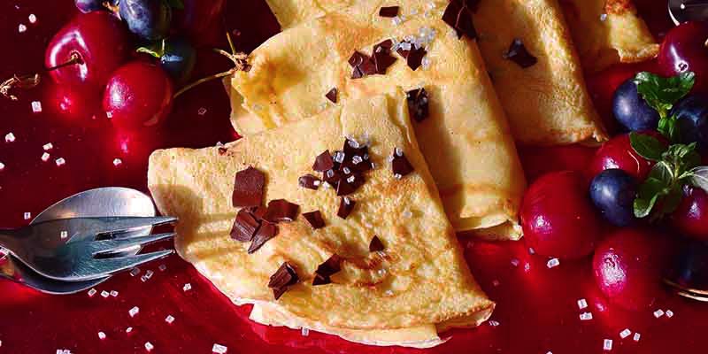 fondo rojo con crepes en fila, las cuales tienen trozos de chocolate y frutos rojos a ambos lados