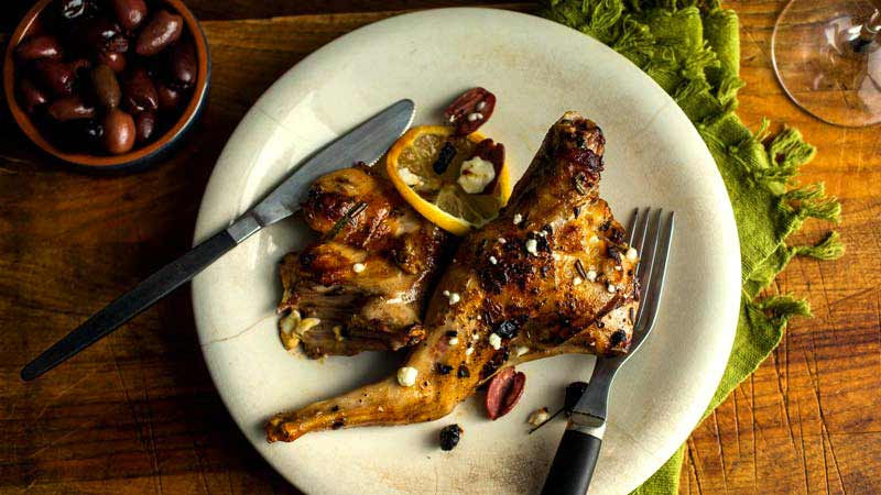 plato blanco con dos piezas de conejo y cubiertos a cada lado, además de estar sobre un mantel verde y tener un recipiente con aceitunas a un lado