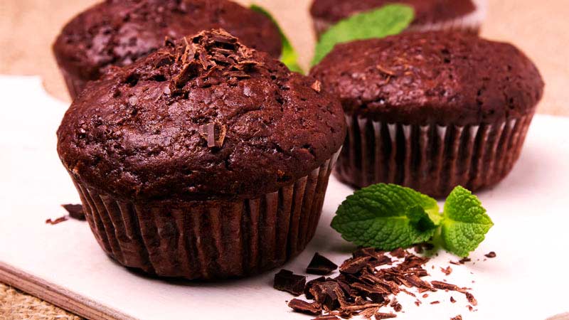 cuatro muffins hechos de chocolate con hojitas verdes y con virutas de chocolate encima y a un lado