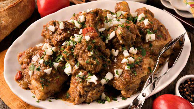 piezas de pollo en una bandeja blanca con cubierto metálico a un lado y rodeado de tomates y pan