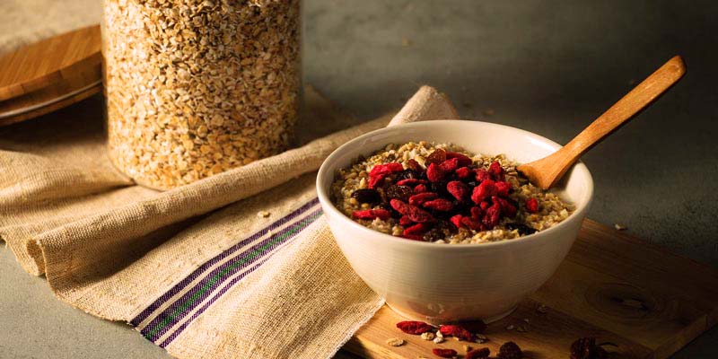 bol blanco con avena y frutos rojos, así como también una cuchara de madera. Aparte, este bol está encima de una tabla de madera y al lado de un mantel y de un tarro con avena