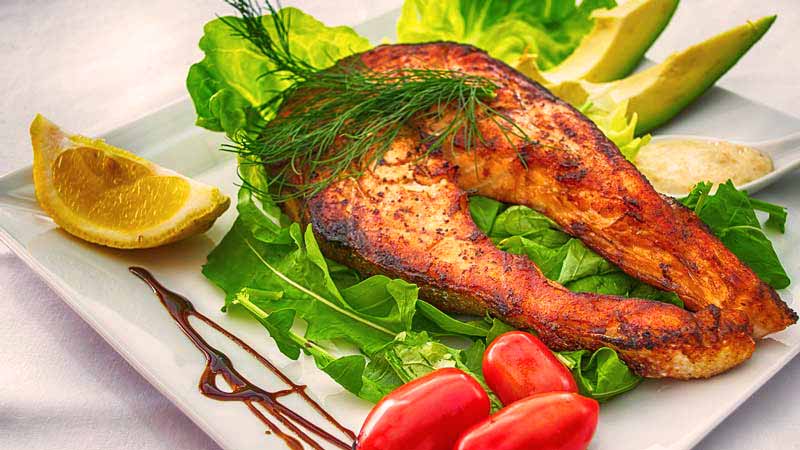 filete de salmón cocinado con lechuga debajo y rodeado de tomates, limón y aguacate