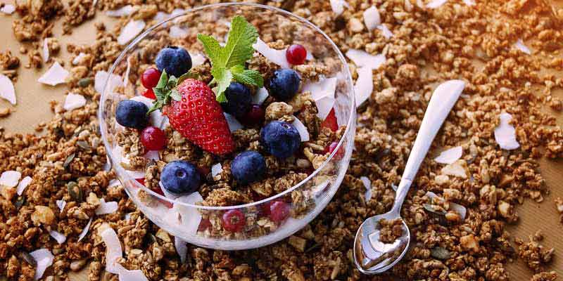 un bol de cristal con yogur, avena y frutos rojos rodeado de más avena y de una cuchara metálica