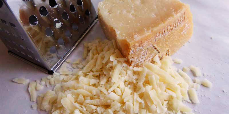 trozo de queso con más queso rallado al lado y con un rallador metálico