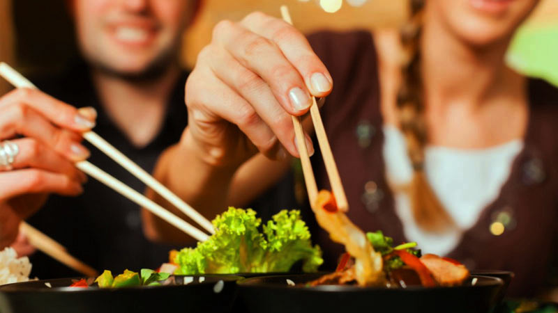 dos personas a las cuales no se les ve la cara y que sostienen unos palillos chinos para coger la comida de unos platos negros