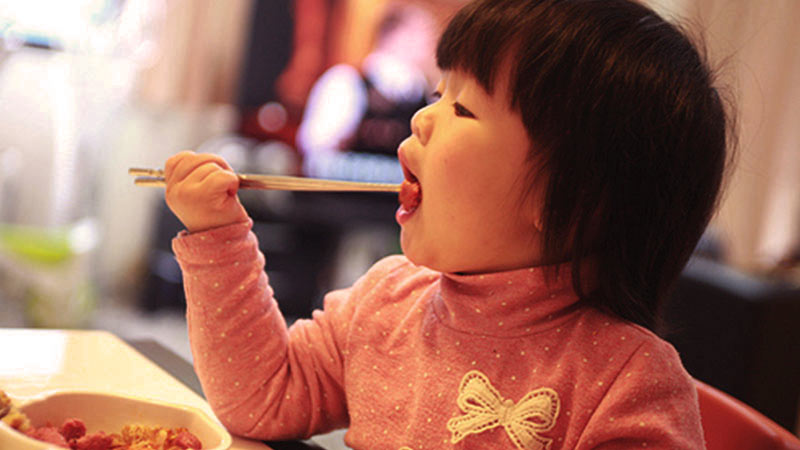 una niña china que se lleva unos palillos chinos a la boca y que tiene un jersey rosa con un lazo blanco