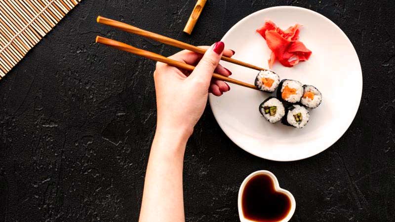 una mano con las uñas pintadas que sostiene unos palillos chinos para coger una pieza de sushi de las cinco que hay en un plato blanco