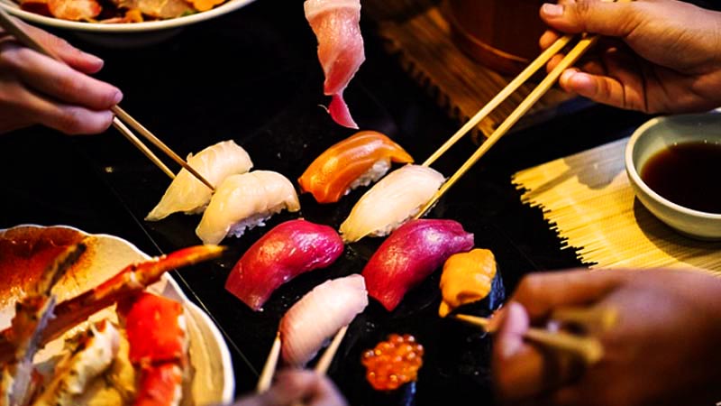 varias manos sosteniendo palillos chinos para coger trozos de sushi que hay en el centro en una placa negra