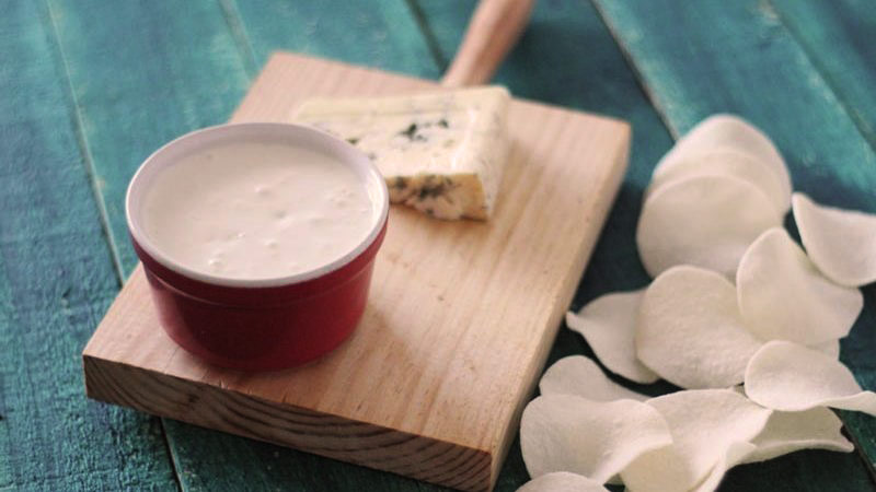 Tabla de madera con queso azul y un bol rojo con salsa, así como también patatas blancas al lado
