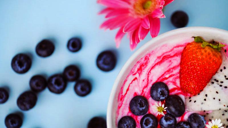 en una esquina, yogur en un bol blanco con frutos rojos y sobre un fondo azul