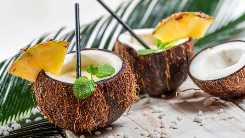 beneficios del coco, cocos con bebida dentro, un trozo de piña en el borde y pajitas negras