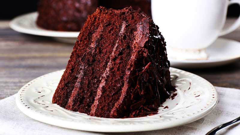 un trozo de bizcocho de chocolate en un plato blanco y al fondo otro trozo y una taza blanca