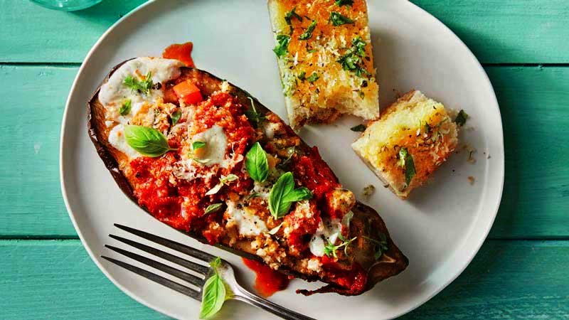 una berenjena rellena en un plato blanco con una rebanada de pan al lado, un tener y sobre un fondo de madera azul